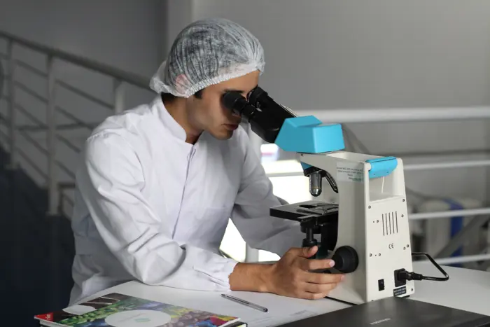 A scientist in a lab coat and hair net conducts testing with a microscope in a laboratory setting. A notebook and pen are visible on the table, along with a magazine featuring colorful images.