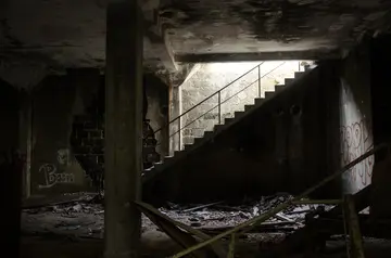 A dimly lit, abandoned building with a concrete staircase leading up. The walls are dark and worn, with visible graffiti. Debris and mold cover the floor, creating an eerie and neglected atmosphere. Light seeps through a gap at the top of the stairs.