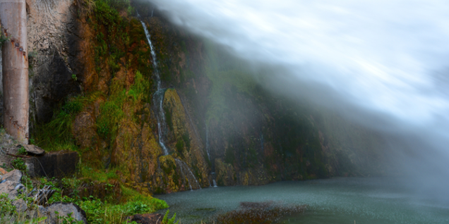 A waterfall cascades down a rocky cliff, partially obscured by mist, into a pool below. Greenery clings to the rocks, offering a lush touch to the serene landscape where nature's beauty invites endless investigation.