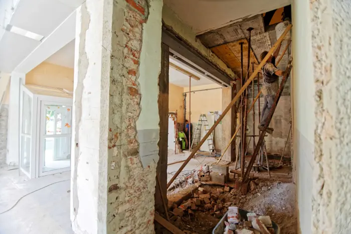 Construction site with exposed brick walls and unfinished surfaces. A worker stands on scaffolding, surrounded by debris, conducting an assessment of the renovation progress. Ladders and tools are visible in the background, indicating ongoing building work.