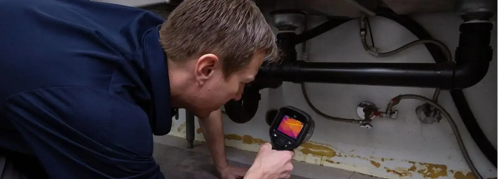 A person conducting a detailed inspection of plumbing pipes under a sink with a thermal imaging camera. Focused on the screen's heat map, they carry out an assessment amidst the industrial surroundings of pipes and fittings.