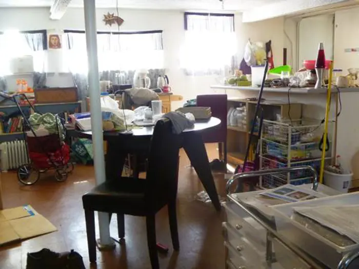 The room is a chaotic mix, featuring a round black table surrounded by chairs, cluttered with clothes and kitchenware. A stroller rests on the left, and shelves are loaded with household goods. Sunlight filters through sheer curtains, inviting a casual inspection of its disarray.