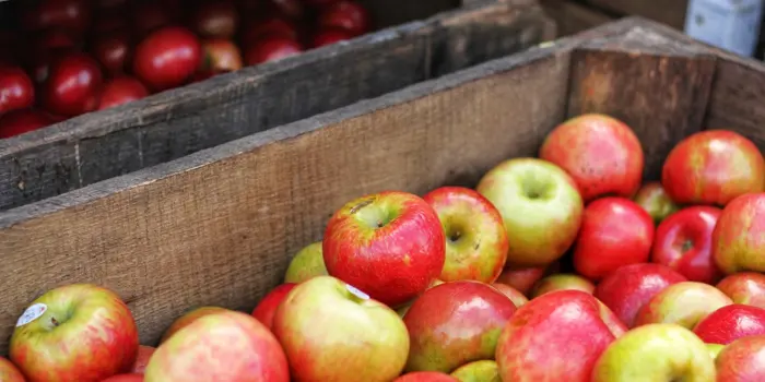 A wooden crate filled with red and green apples is displayed in a market setting, inviting inspection. Another crate, partially visible, contains more red apples in the background.
