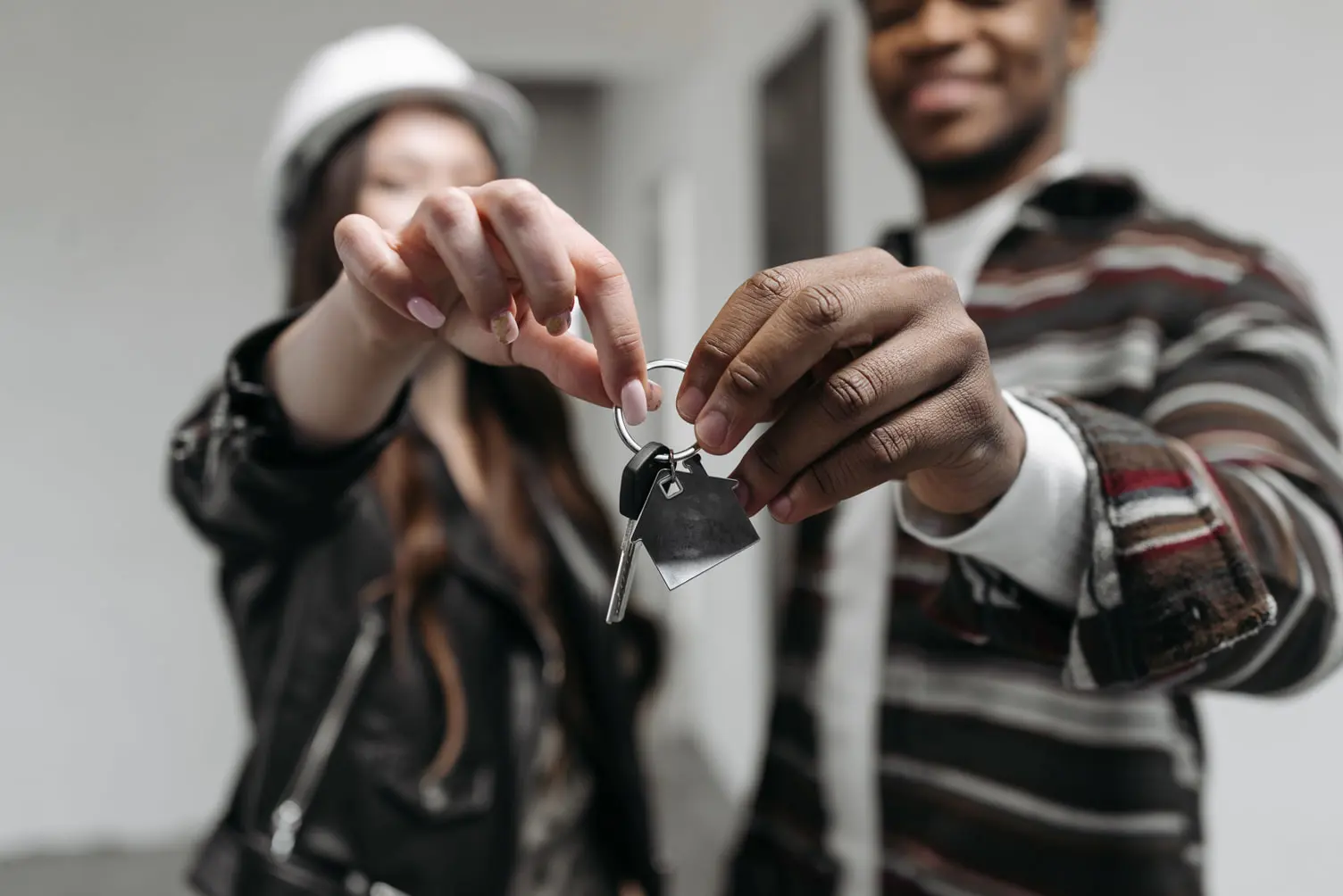 A man and a woman hold a single key together, smiling. The woman wears a hard hat and leather jacket, while the man sports a striped shirt. They stand in an interior space, possibly signifying ownership or the conclusion of an investigative assessment for their new beginning.