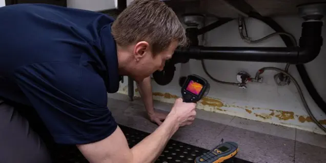 A person uses a thermal imaging camera to inspect plumbing under a sink for any potential mold issues. The camera screen shows an infrared image. Nearby, there is an electronic device with buttons, hinting at further sampling or inspection tasks in this indoor maintenance area.