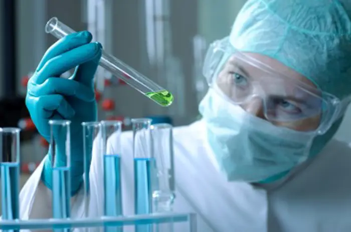 A scientist wearing a mask, goggles, and gloves conducts an investigation of a green liquid in a test tube. Several other test tubes containing blue liquid are in the foreground within the laboratory setting.