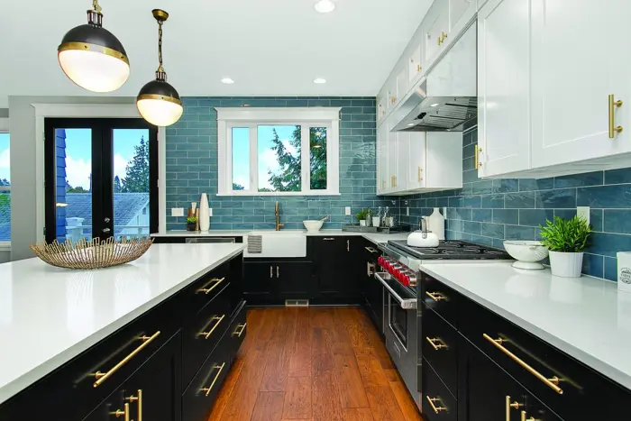 A modern kitchen with black lower cabinets and white upper cabinets features stainless steel appliances, a white countertop, and a teal tile backsplash. Pendant lights hang above an island with a decorative tray, adding flair. A window offers a view of trees, ensuring natural light for mold testing.