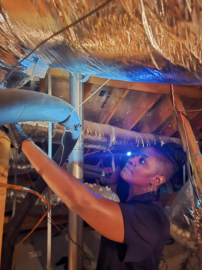 A person is conducting an inspection in an attic, holding a flashlight. The space has exposed wooden beams and reflective insulation. Focused on sampling from a large pipe, the individual wears gloves and a dark shirt, with braided hair tied up.