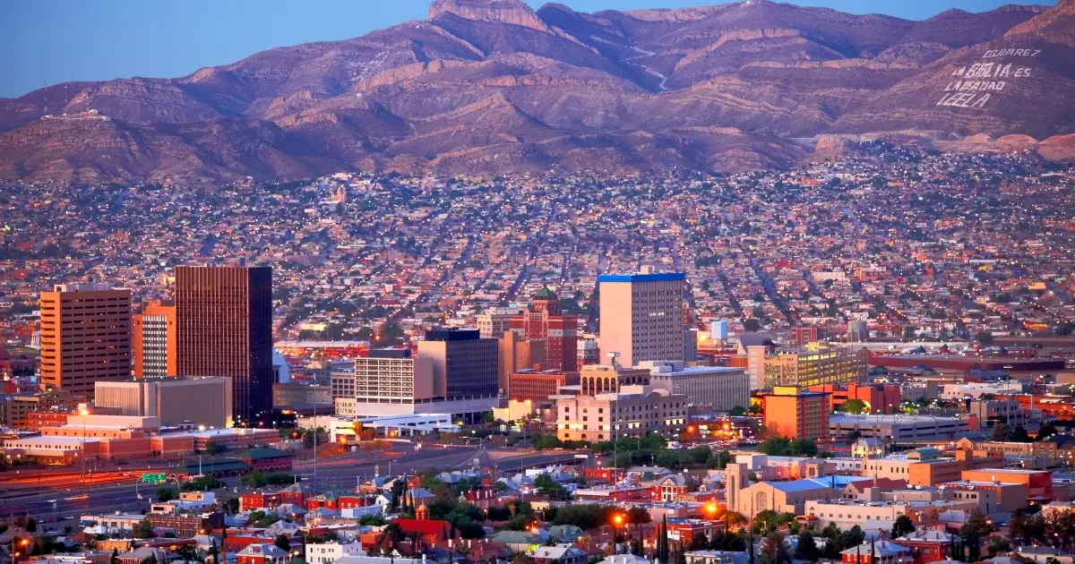 A vibrant cityscape at sunset shows tall buildings in the foreground, spreading into a densely packed residential area, with a mountain range in the background—a scene perfect for the investigation and sampling of diverse urban life.