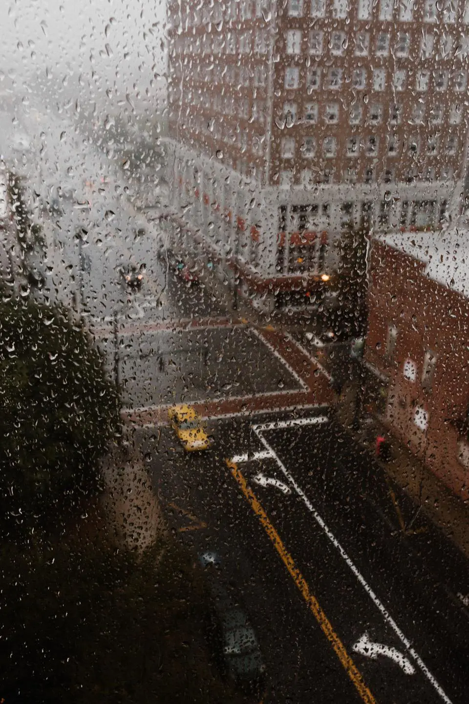 View from above of a rainy city street intersection. Raindrops cover the window, blurring the scene like a filter used in mold inspections. A yellow taxi is visible below, surrounded by wet roads and brick buildings. The atmosphere is overcast and moody.