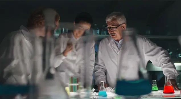 A group of scientists in lab coats are engaged in discussion around a table, conducting an assessment of various colorful liquids in flasks. The dimly lit laboratory accentuates the vibrant hues of the chemicals, creating an atmosphere ripe for discovery.