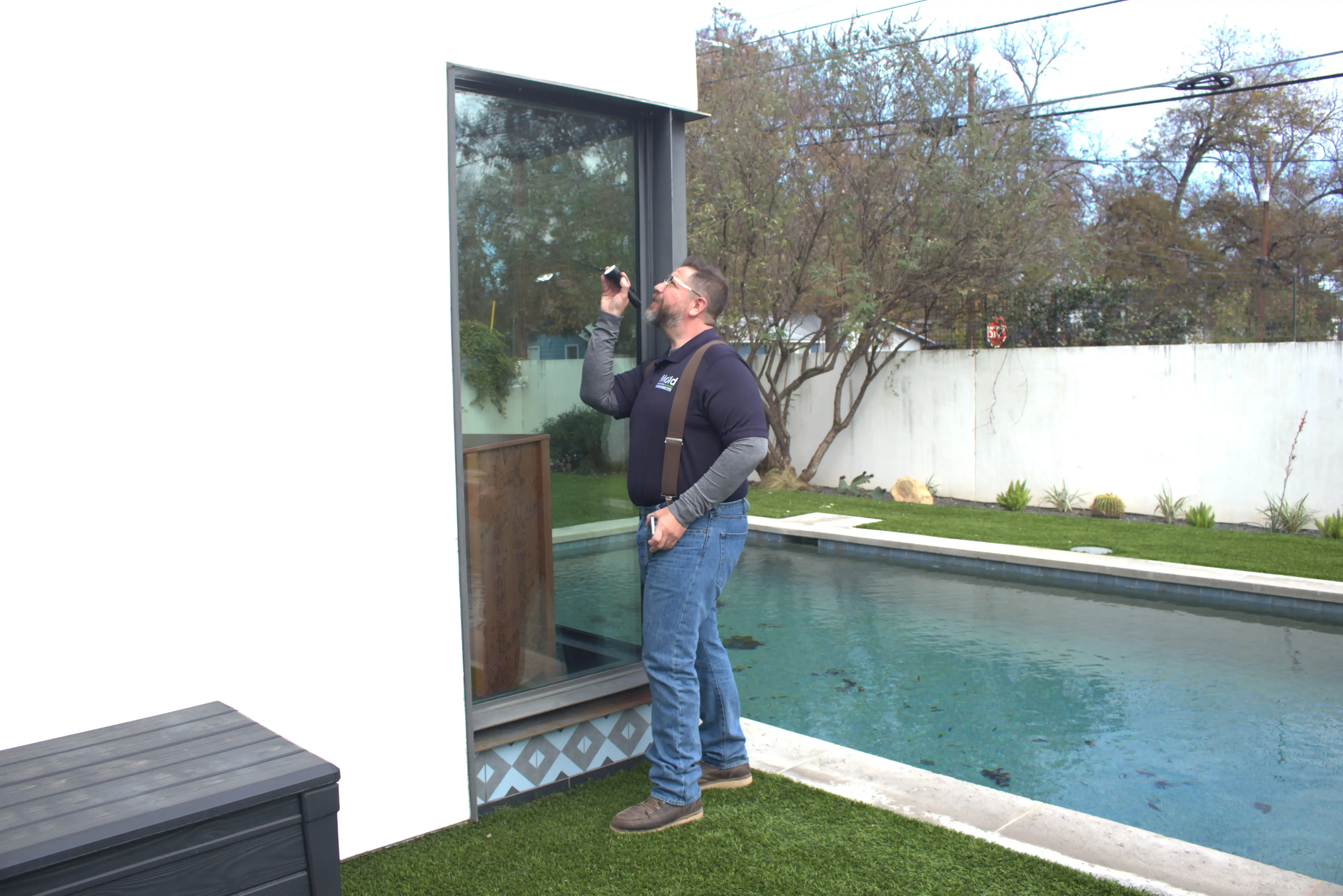 A man in a navy blue shirt and jeans inspects a large window using a flashlight, likely conducting an investigation. He stands beside a modern house with a swimming pool and neatly trimmed lawn in the background, searching for any signs of mold or other issues.