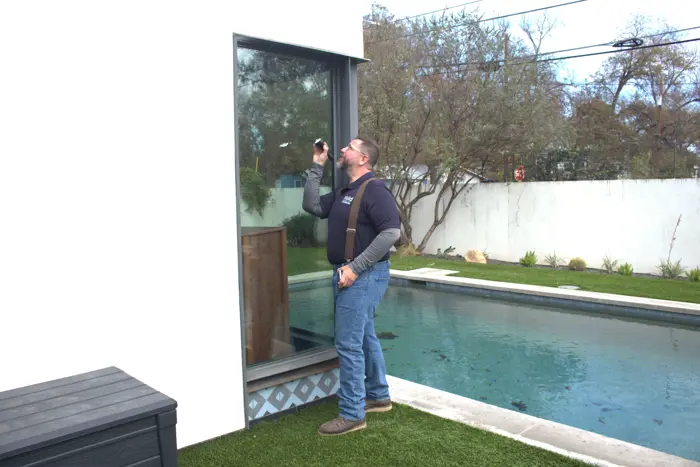 A man in a navy blue shirt and jeans inspects a large window using a flashlight, likely conducting an investigation. He stands beside a modern house with a swimming pool and neatly trimmed lawn in the background, searching for any signs of mold or other issues.