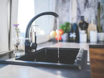 A modern kitchen sink with a black faucet is shown, water gracefully flowing, as if sampling the serene ambiance. Beside the sink rests a small glass vase holding a purple and yellow flower. In the background, blurred kitchen utensils and a cutting board complete the elegant scene.