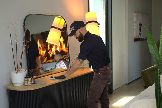A bearded man wearing a cap and gloves conducts an inspection as he cleans a modern console table by the lit fireplace. The room, featuring a large mirror, decorative objects, and plants, creates a cozy atmosphere.